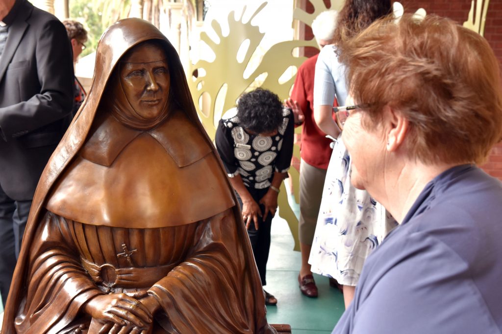 A prayerful and engaging woman, Ursula Frayne is depicted sitting on a steamer trunk with her hands resting on her breviary. She looks at the viewer who may choose to sit on the opposite suitcases, inviting them to commute with her. Photo: Feby Plando