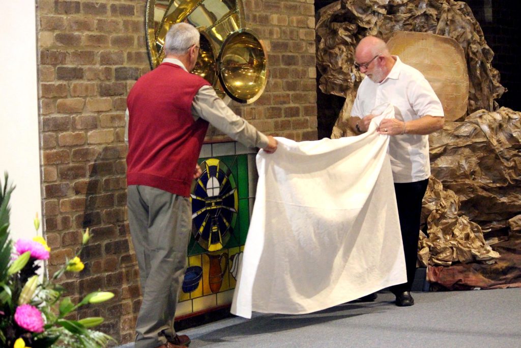 John Griffin and Alan Kitto unveiling a jarrah-encased, stained glass panel that they crafted and donated to the parish. The panel showcases pictures relating to the Eucharist and the washing of the feet. It will highlight the tabernacle area in the church and bears special relevance to the spiritual journey of the Somascan Order. Photo: Supplied